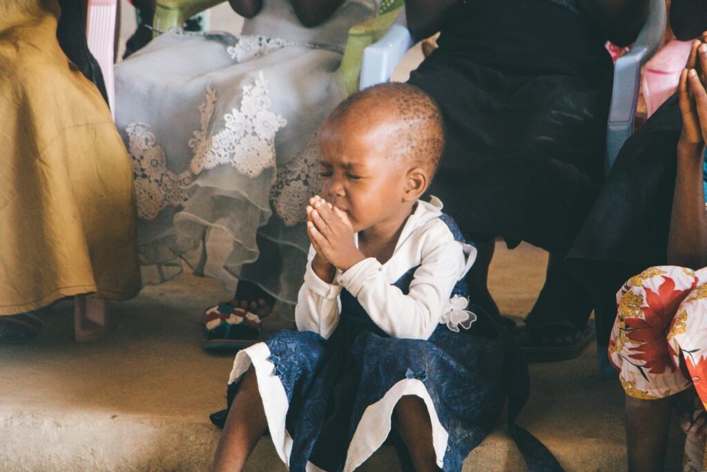 A young child in Tanzania prays in contemplation during a community gathering, symbolizing faith and innocence.